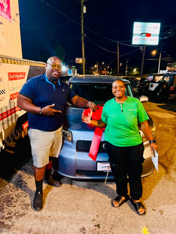 TQ standing with a smiling customer beside a vehicle with a large red bow outside First Class Auto Sales at night