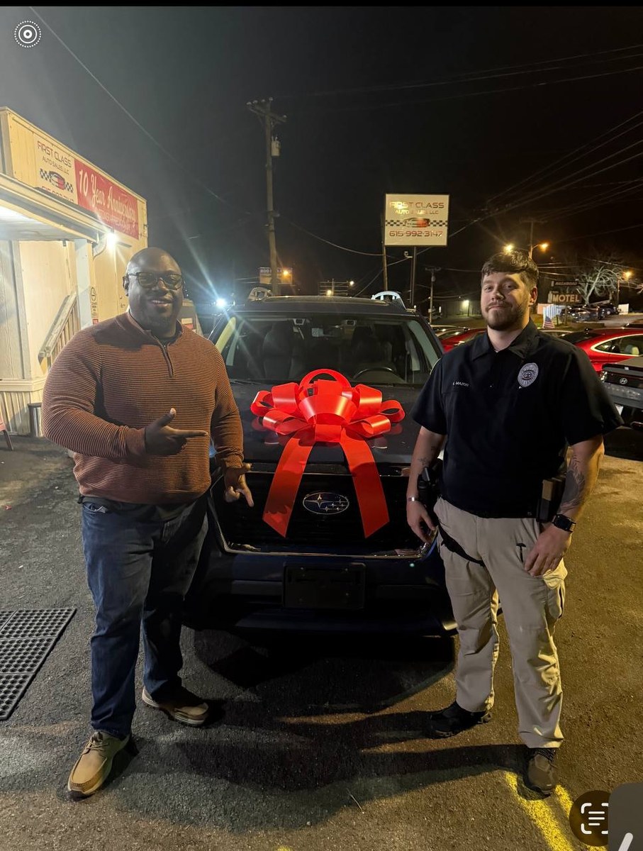 TQ standing with a customer beside a Subaru SUV with a large red bow at First Class Auto Sales at night